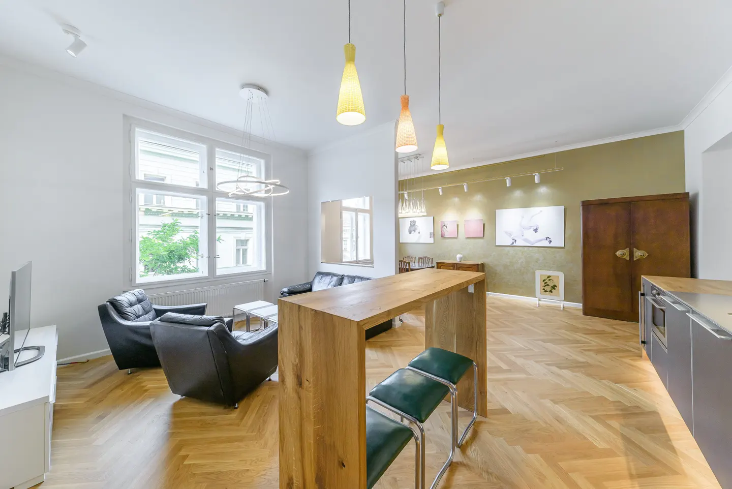 Bright, open apartment with wood floors, black leather chairs, and a wooden bar with green stools. Artwork adorns the olive-green wall.