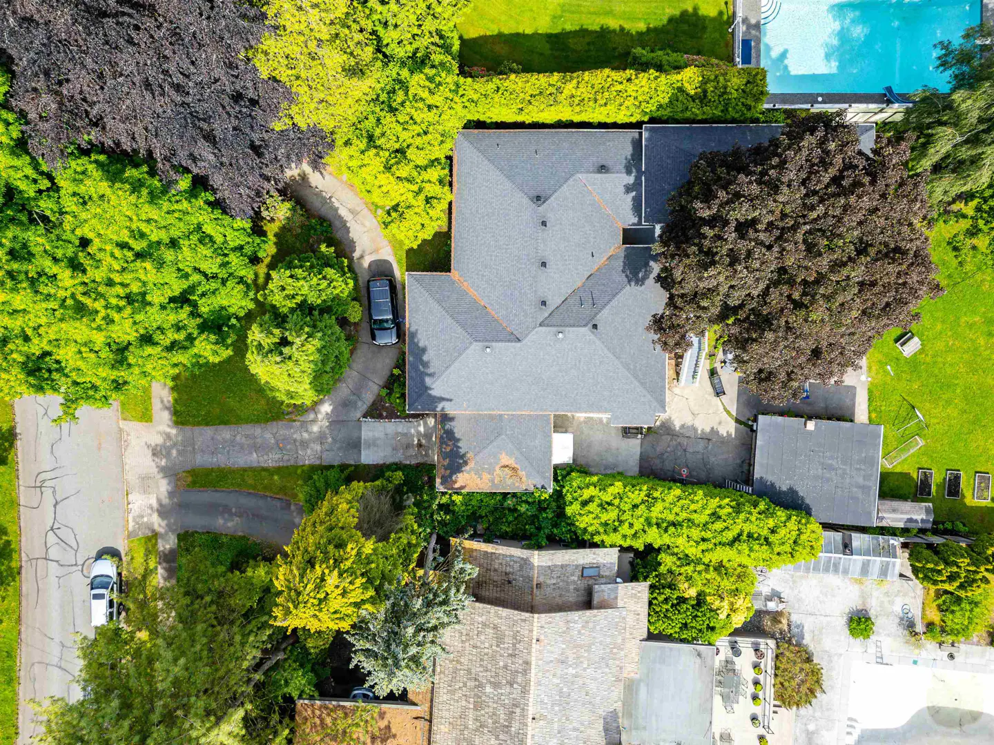 Aerial view of a gray roofed house surrounded by green trees, a pool, and a driveway with a car.