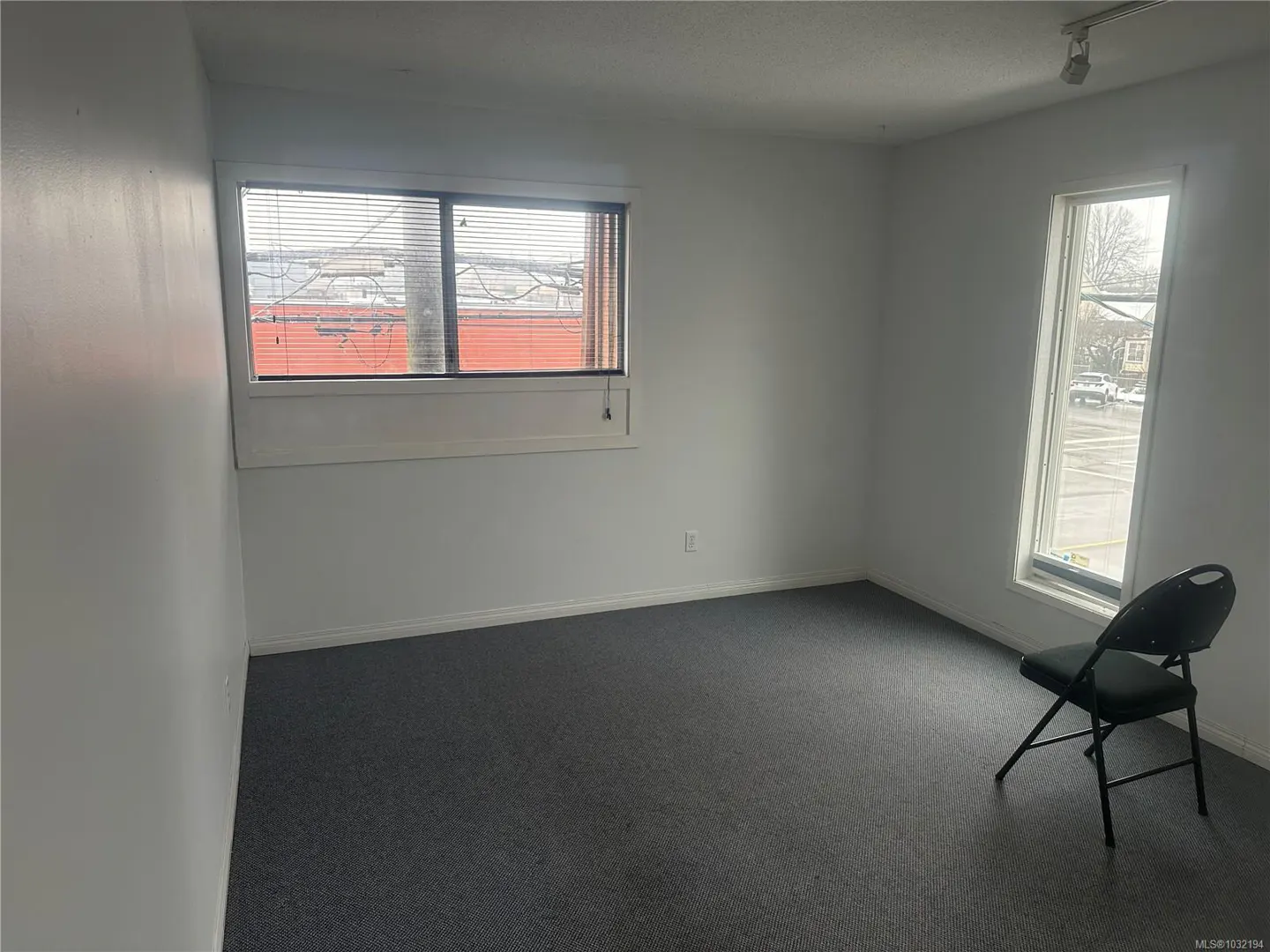 A vacant room with white walls, gray carpet, and two windows. A black folding chair sits near the right window.