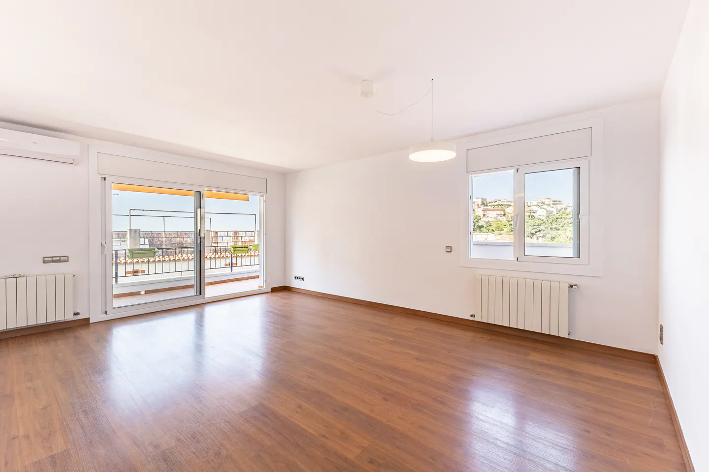 Bright, empty room with wood floors, white walls, and a balcony view. A window shows a cityscape. A modern light fixture hangs from the ceiling.