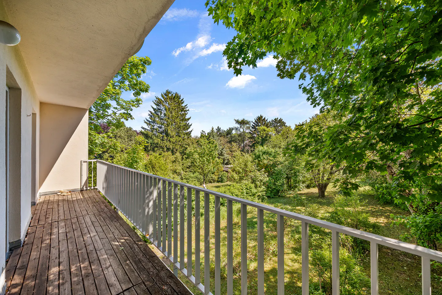 Balcony with wooden floor and white railing overlooking a lush green garden under a blue sky.