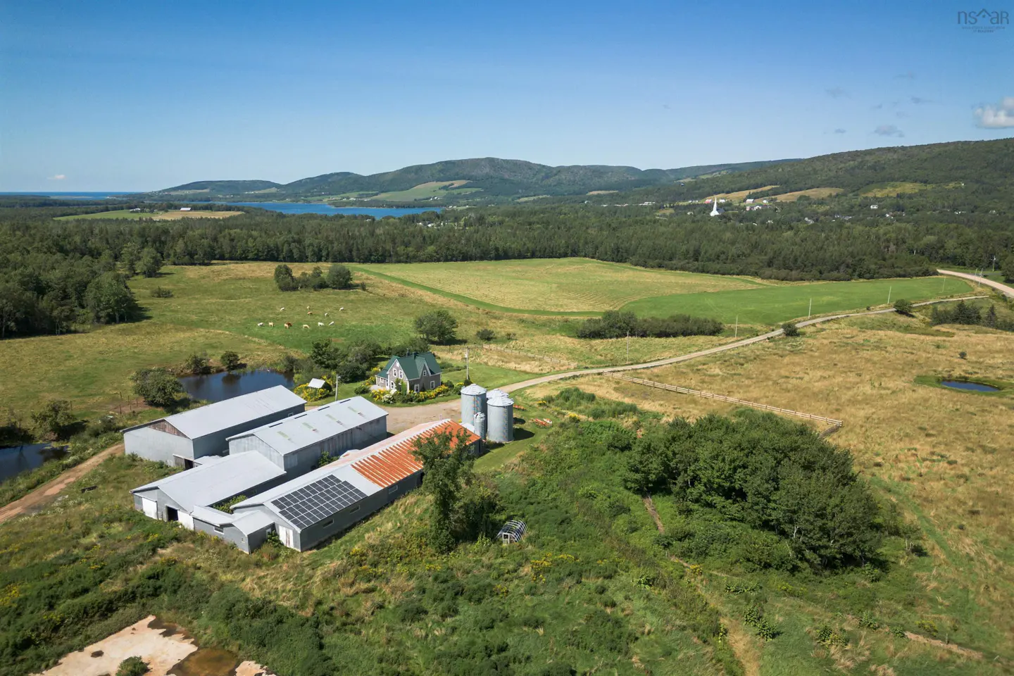 Aerial view of a farm with barns, silos, and solar panels in a green, rural landscape under a blue sky.