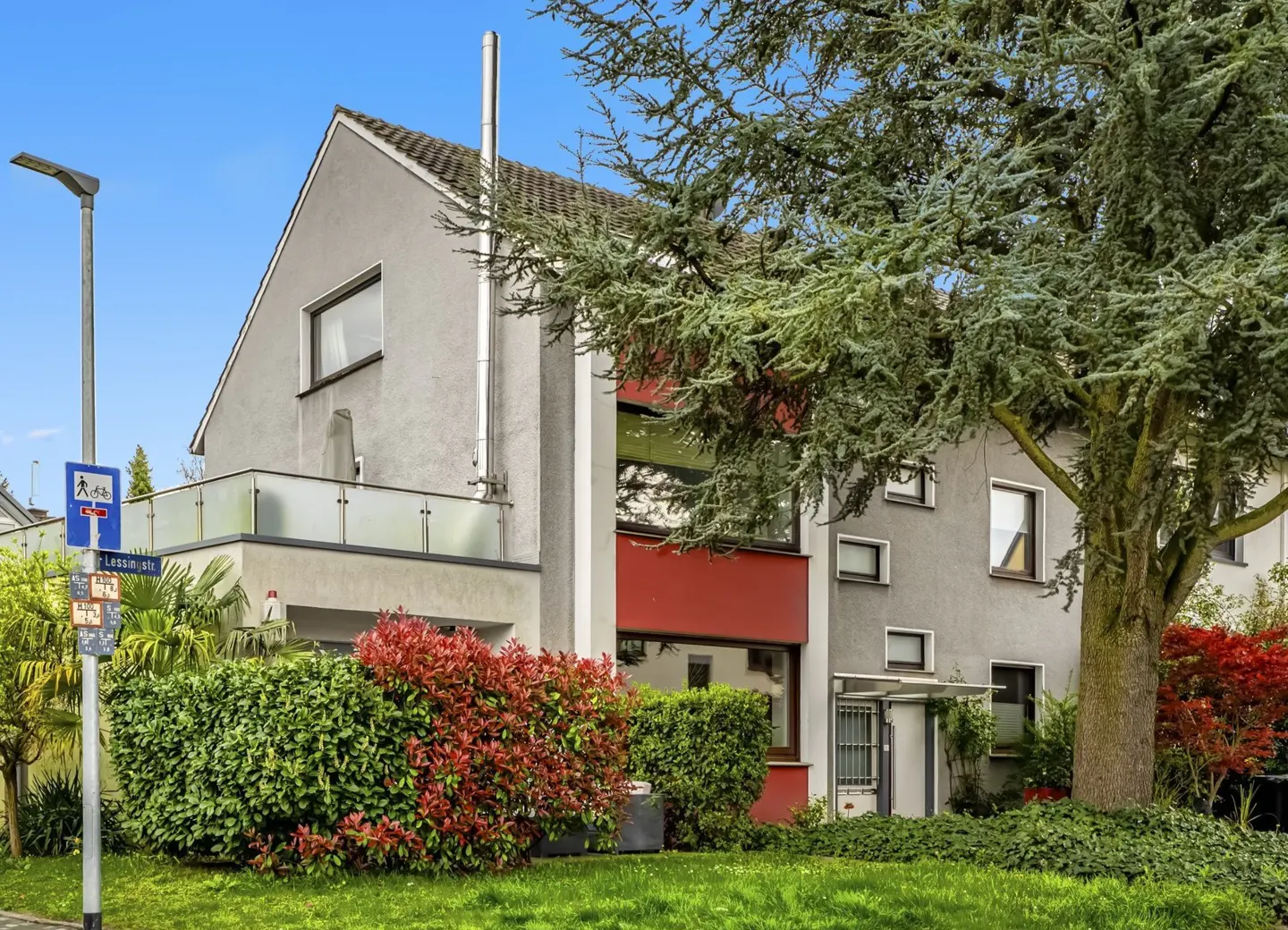 A modern gray house with a red accent wall, a balcony, and a metal chimney, surrounded by green bushes and a large tree.