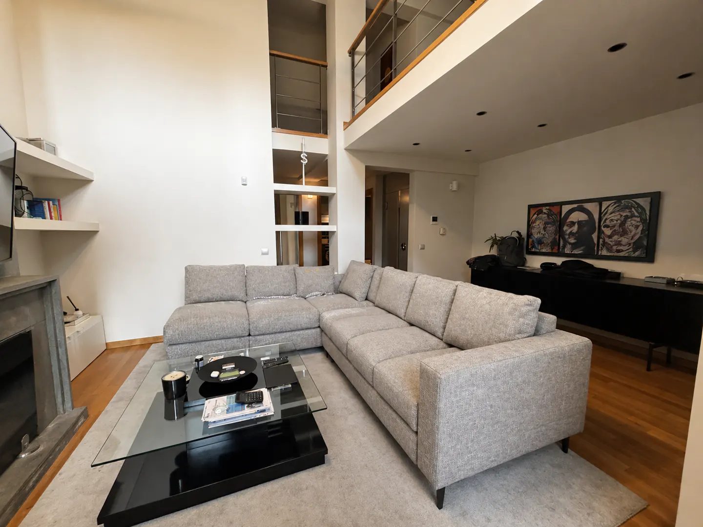 A modern living room with a gray sectional sofa, a glass coffee table, and a two-story wall with a balcony.