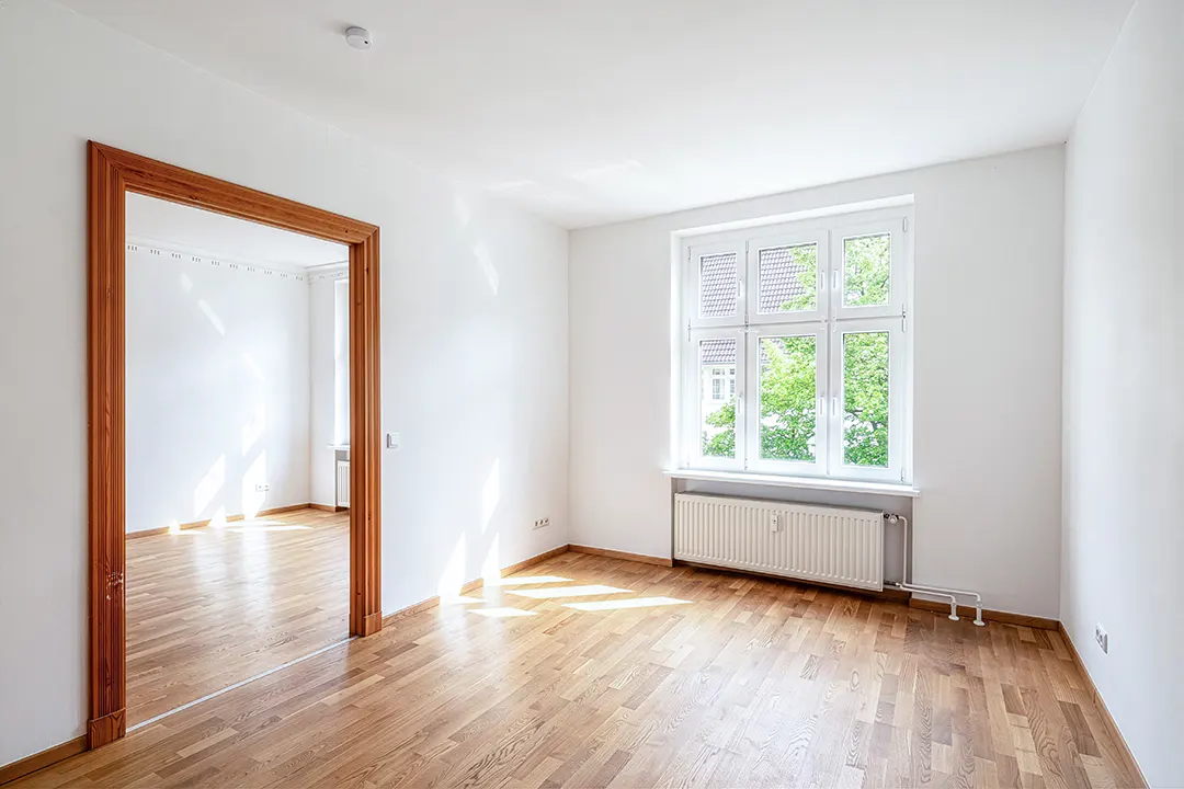 Bright, empty room with hardwood floors, white walls, and a large window. A wooden door frame leads to another room.