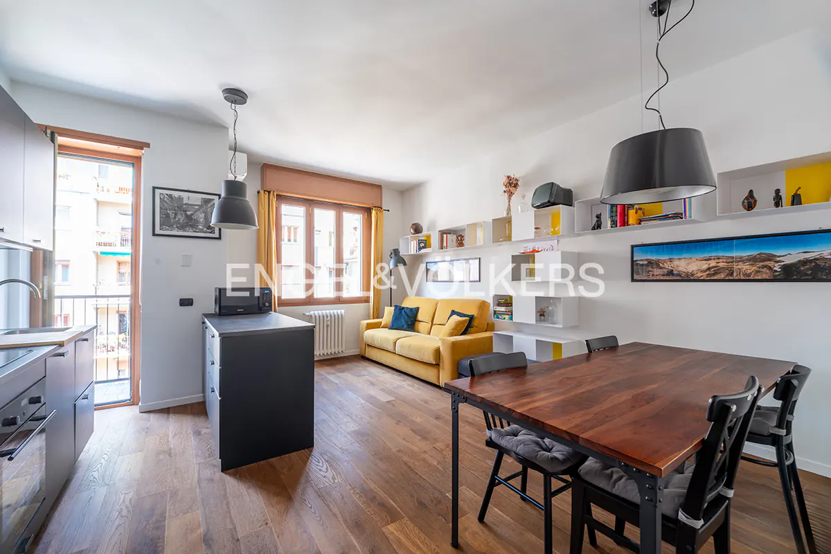 Bright apartment interior with wood floors, a yellow sofa, and a dark wood dining table with black chairs. Shelves line the wall.