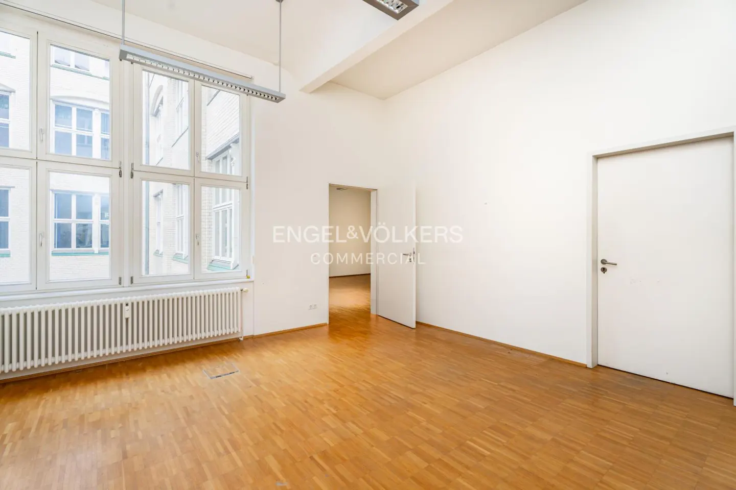 Bright, empty office with light wood floors, white walls, large windows, a radiator, and two white doors.