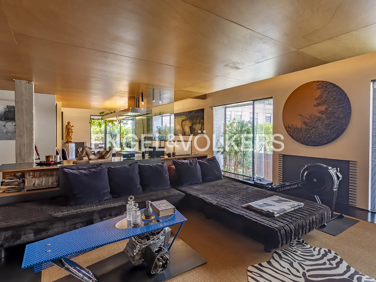 Modern living room with a large black sectional, blue metal coffee table, rowing machine, and a circular wall art piece.