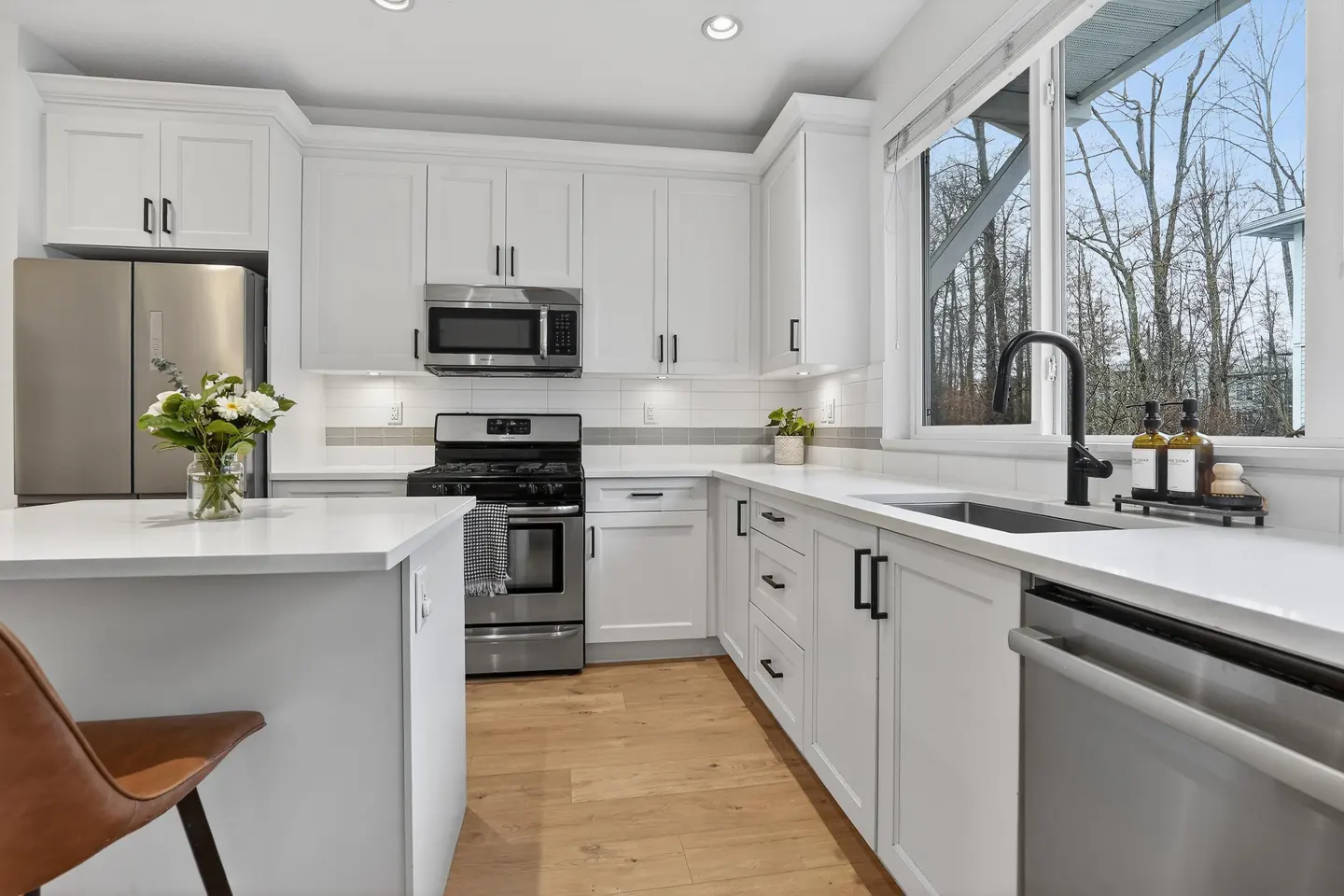 Bright kitchen with white cabinets, stainless steel appliances, and wood floors. A window overlooks trees.