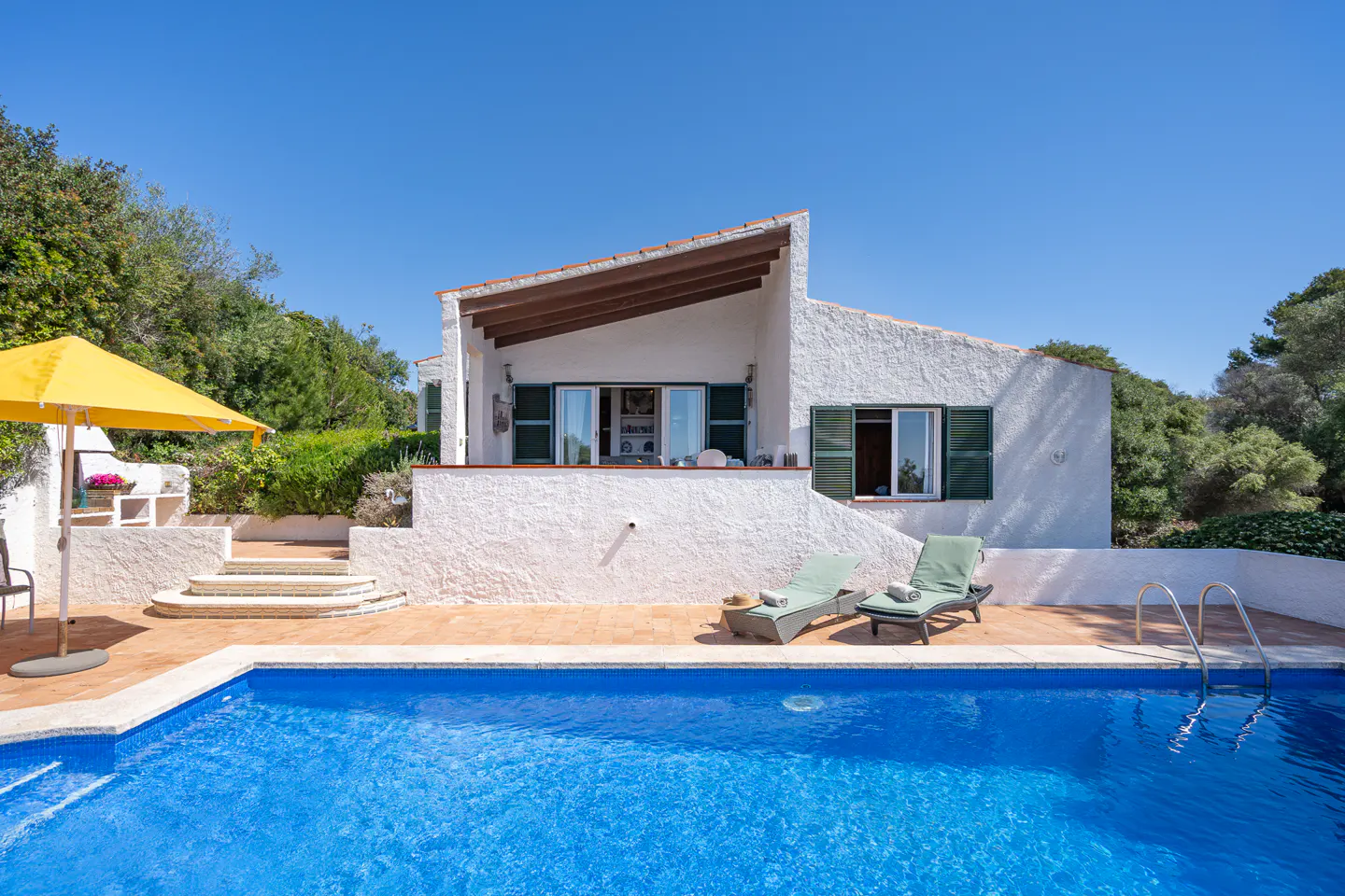 Exterior of a white stucco house with a blue pool, green shutters, and a yellow umbrella.