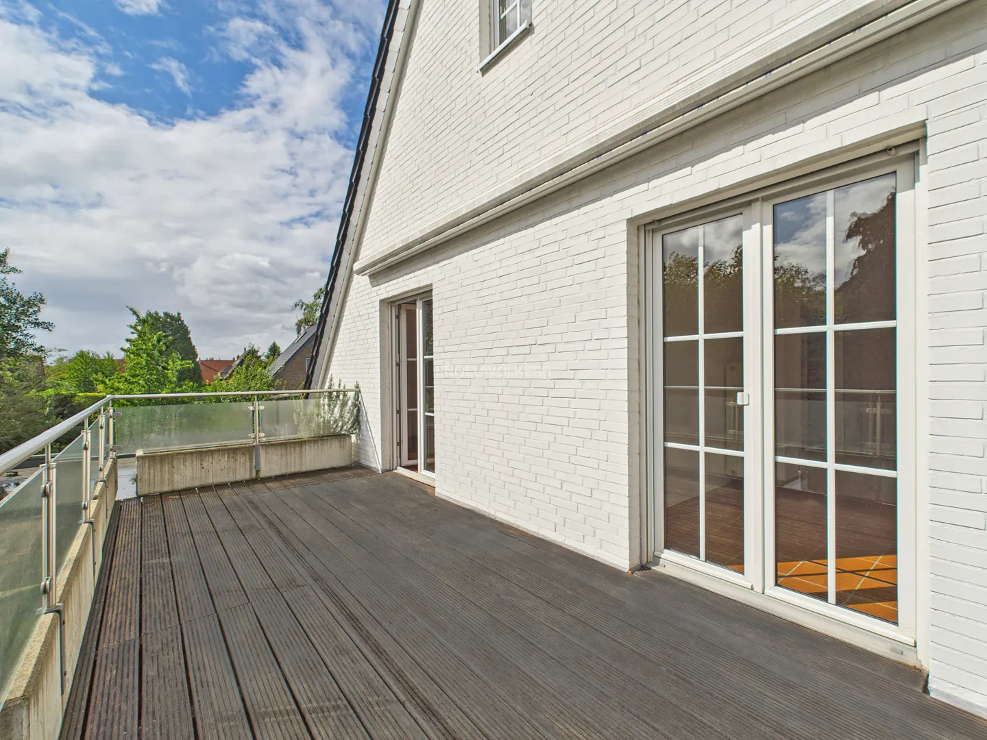 Exterior view of a house with a wooden deck, white brick wall, and glass railing under a cloudy sky.