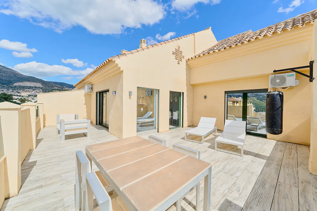 Outdoor patio with white furniture, a dining table, and lounge chairs. A punching bag hangs on the wall. The building is beige with a tiled roof. Blue sky.