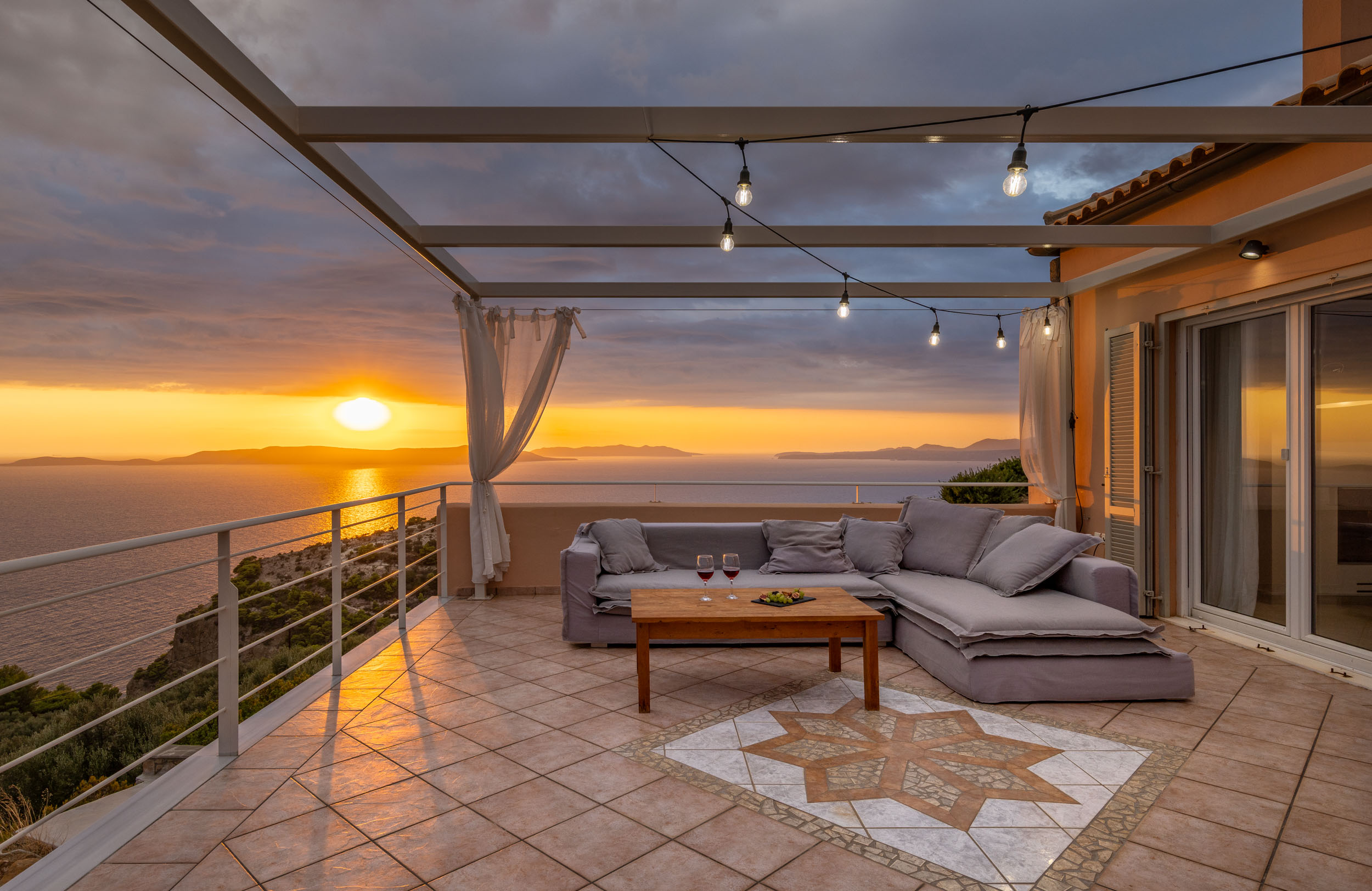 Sunset view from a tiled patio with a gray sofa, wooden table, and string lights. The sun reflects on the ocean.