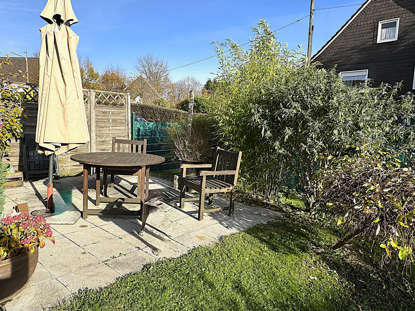 Outdoor patio with a round wooden table, chairs, and a closed beige umbrella. Green lawn and bushes surround the patio.