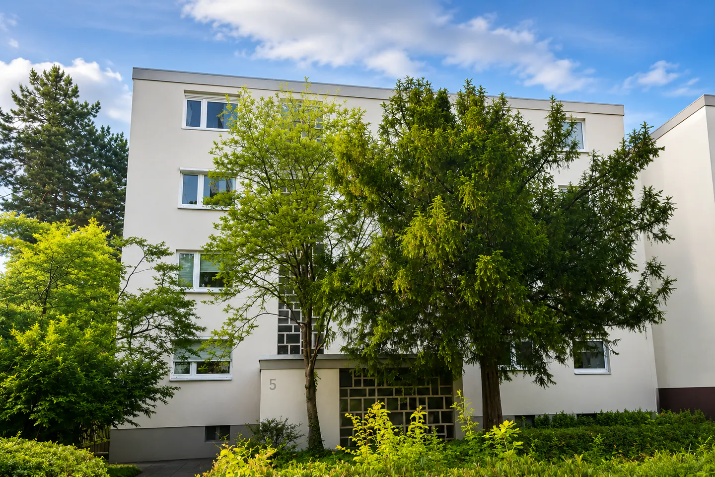 A four-story white apartment building with the number 5, surrounded by lush green trees and bushes under a blue sky.