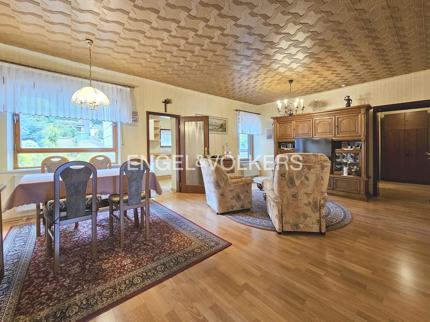 A living and dining area with wood floors, patterned ceiling, dining table with chairs, and floral armchairs.