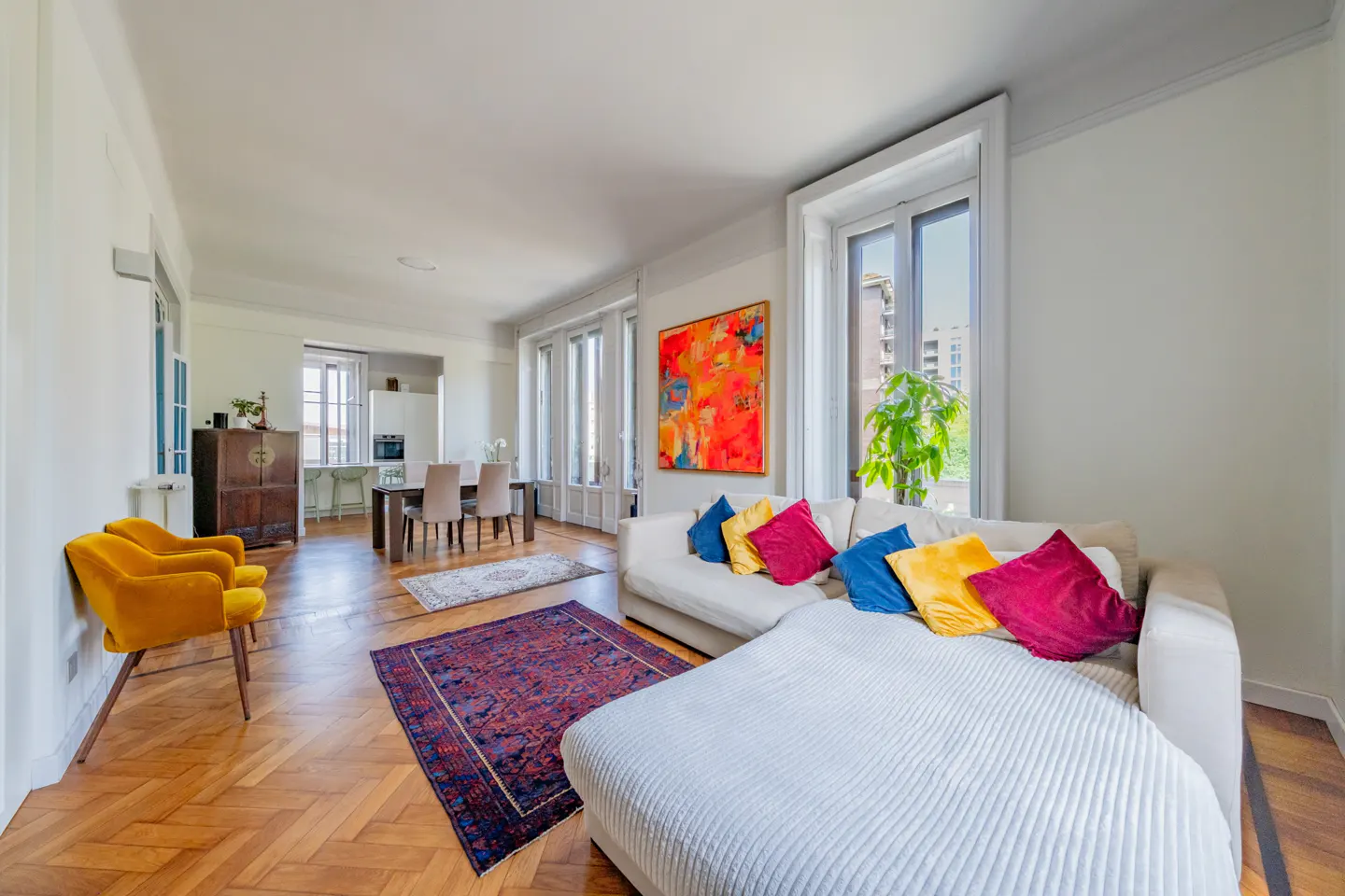 Bright, airy living room with hardwood floors, white walls, and large windows. A white sofa with colorful pillows sits near an abstract painting.