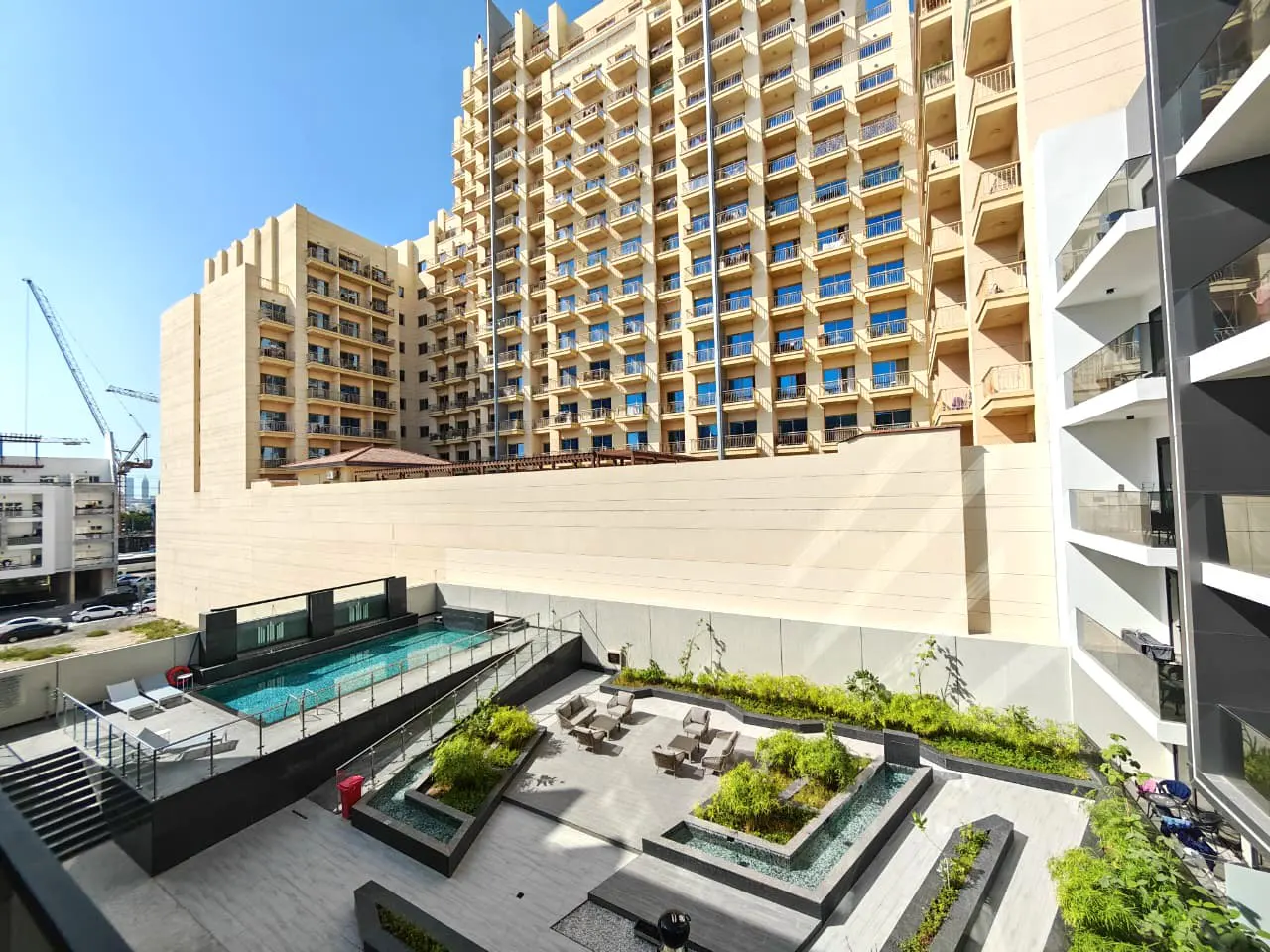 View of a building complex with a pool, lounge area, and greenery on a sunny day.