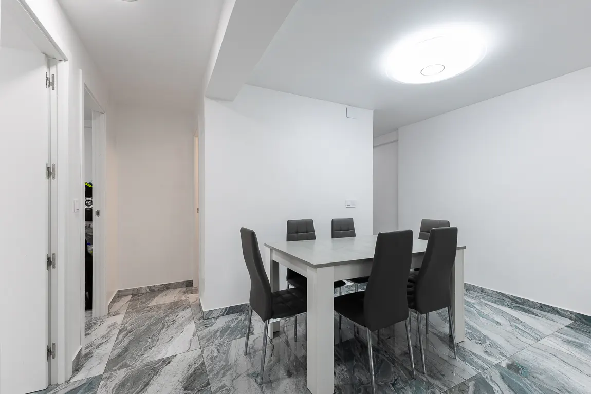 A bright, modern dining room with a white table, six gray chairs, and marble-patterned tile flooring. A doorway is visible on the left.