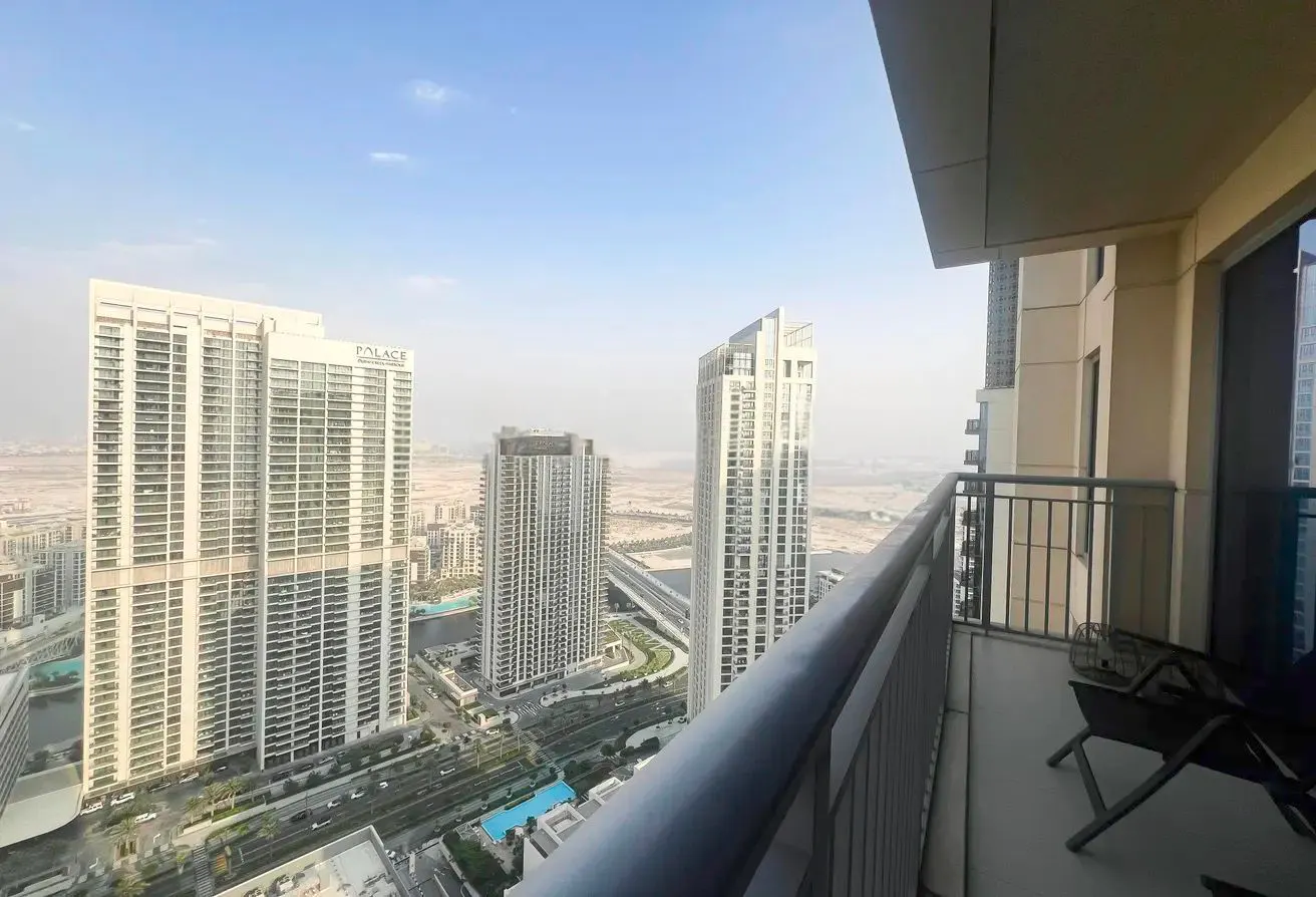 Balcony view of Dubai skyline with tall buildings. Metal railing and chairs on the balcony. Blue sky.
