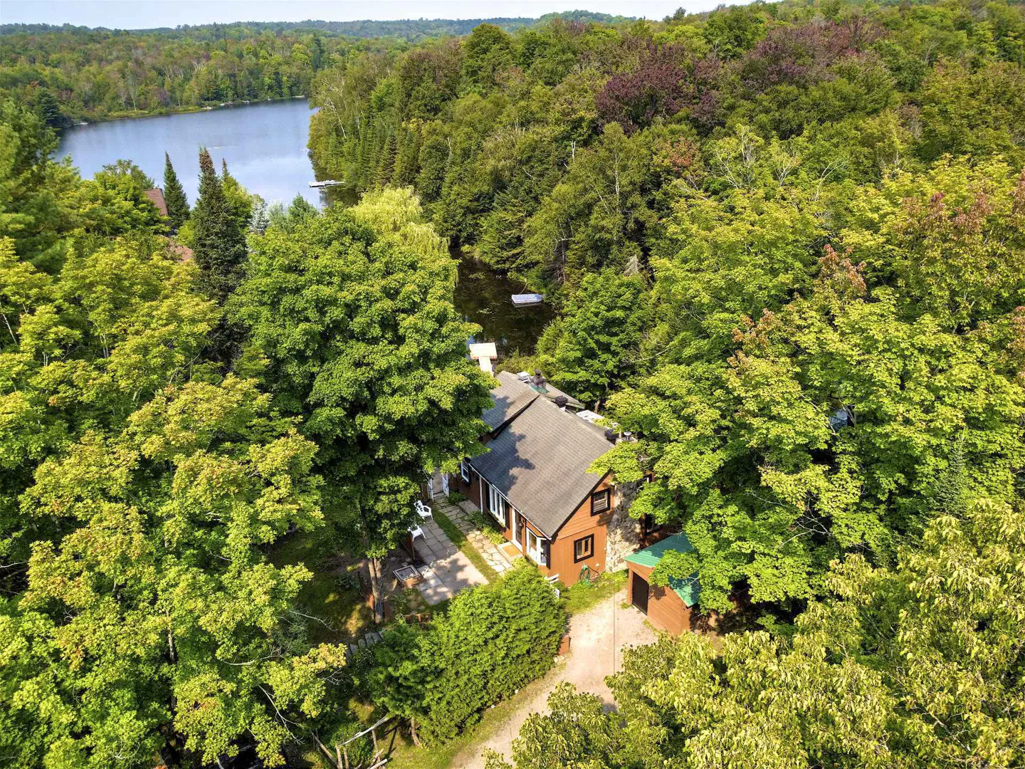 Aerial view of a brown house with a gray roof surrounded by green trees and a lake in the background.