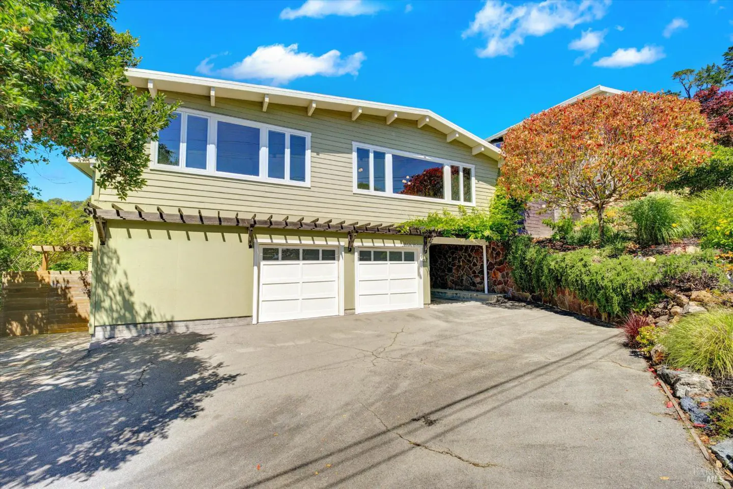 Two-story light green house with white trim, two-car garage, and a stone-covered carport. Landscaped yard with trees and blue sky.