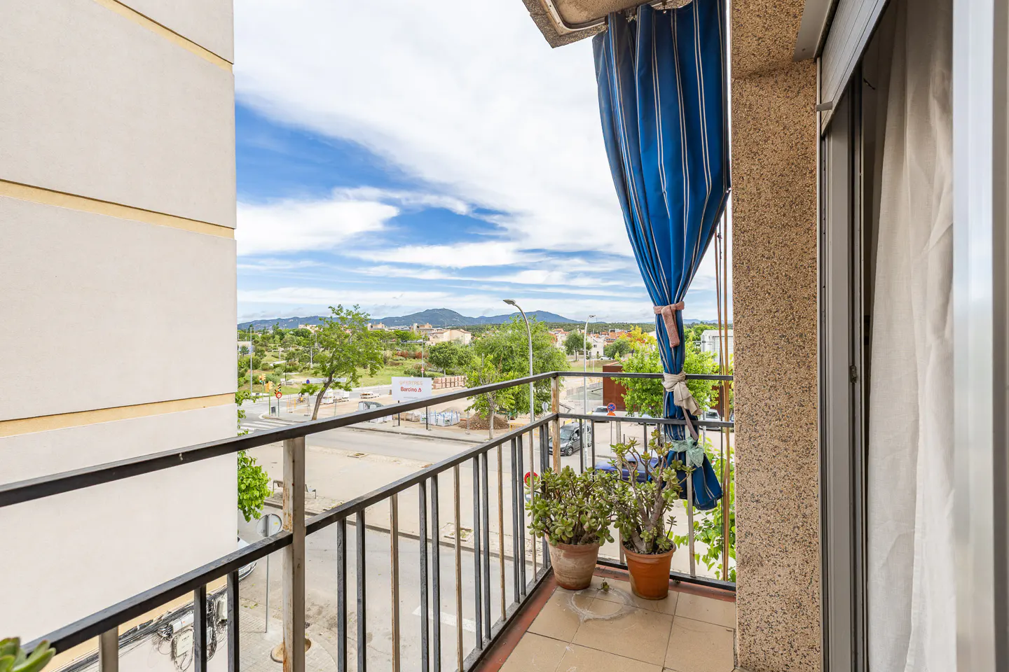 Balcony view with black railings, potted plants, and a blue striped curtain. Cityscape and cloudy sky visible in the background.