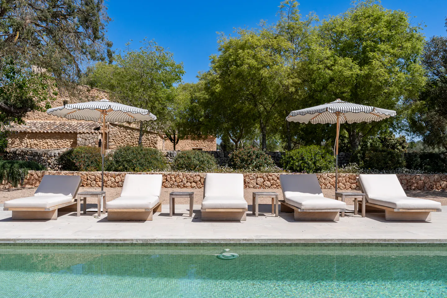 Outdoor pool area with six lounge chairs, small tables, and striped umbrellas. Stone wall and green trees in the background.