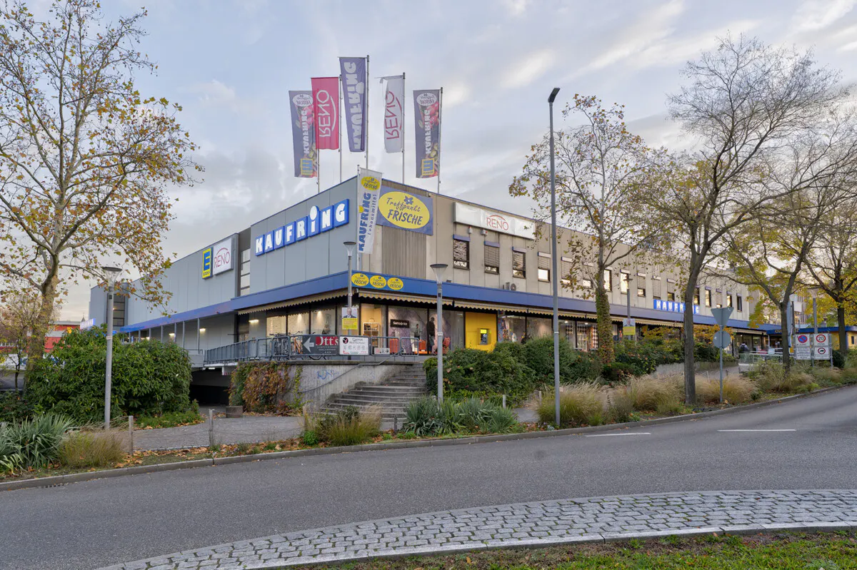 Exterior view of a Kaufring shopping center with flags, trees, and a street in front.