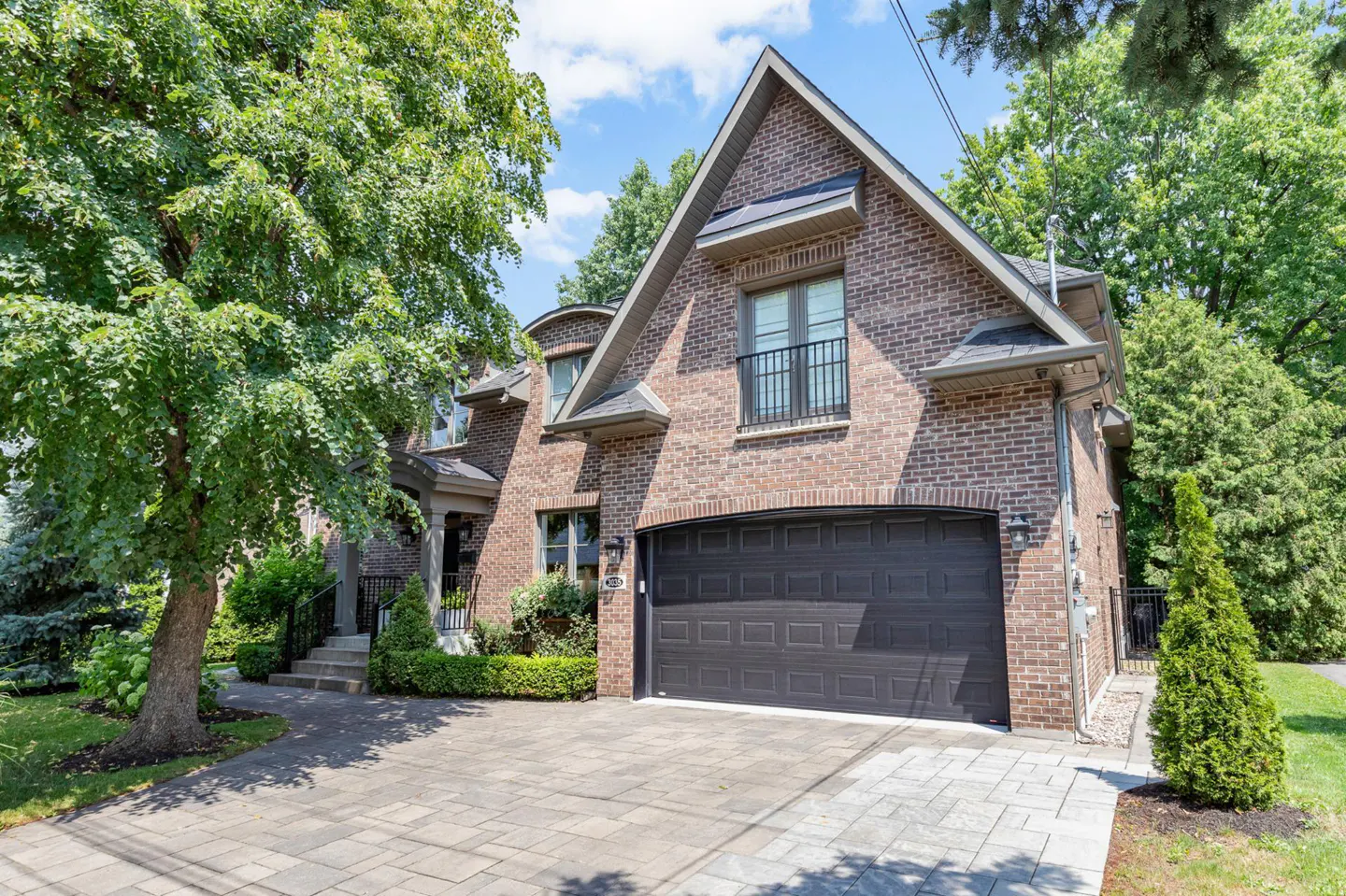 Two-story brick house with a black garage door and a small balcony, surrounded by green trees.
