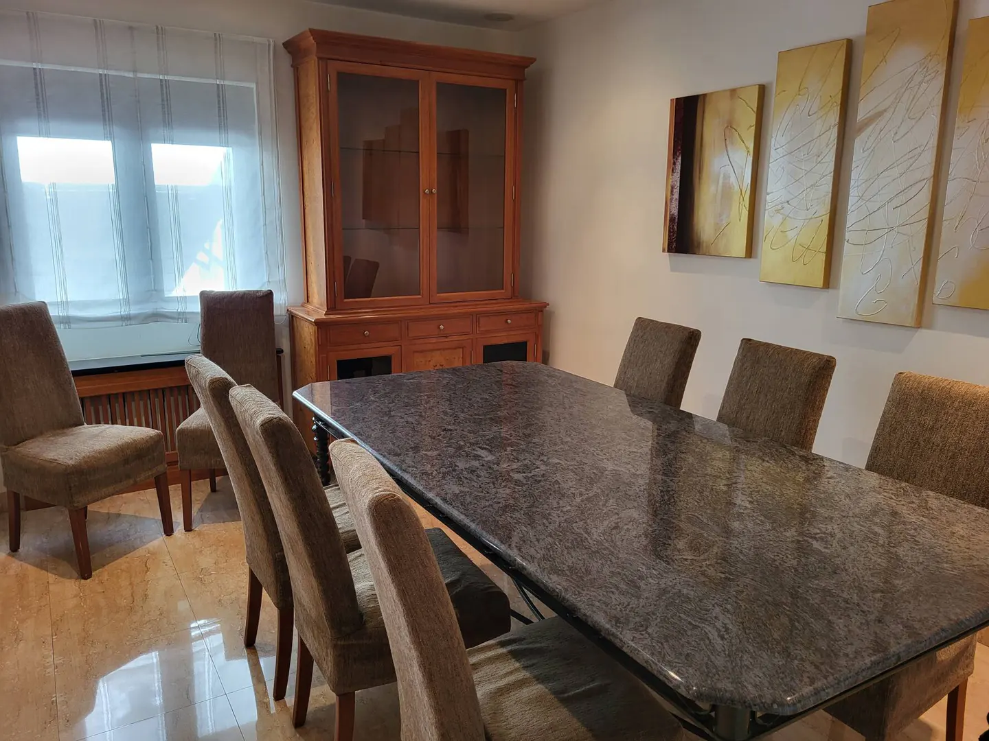 A dining room with a large granite table, brown chairs, and a wooden cabinet. Abstract art hangs on the wall.