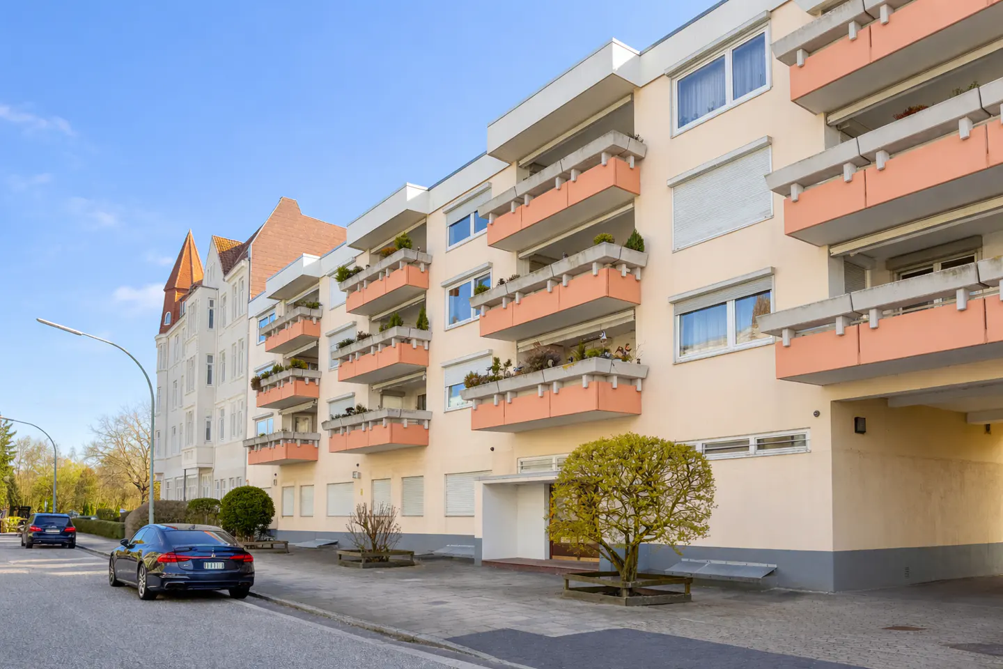 Apartment building with peach balconies and white trim, next to a white building with a red roof. Cars are parked on the street.