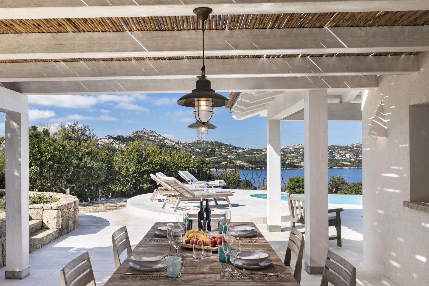 Outdoor dining area with a wooden table set for a meal, overlooking a pool and ocean view. White pergola with hanging lights above.