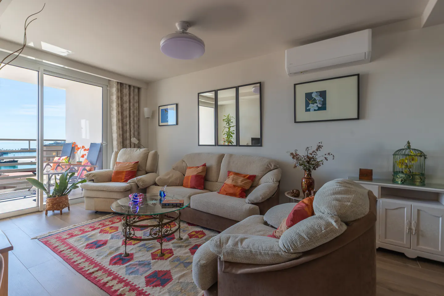 Bright living room with beige sofa, armchairs, and colorful rug. Balcony with ocean view visible through sliding glass doors.