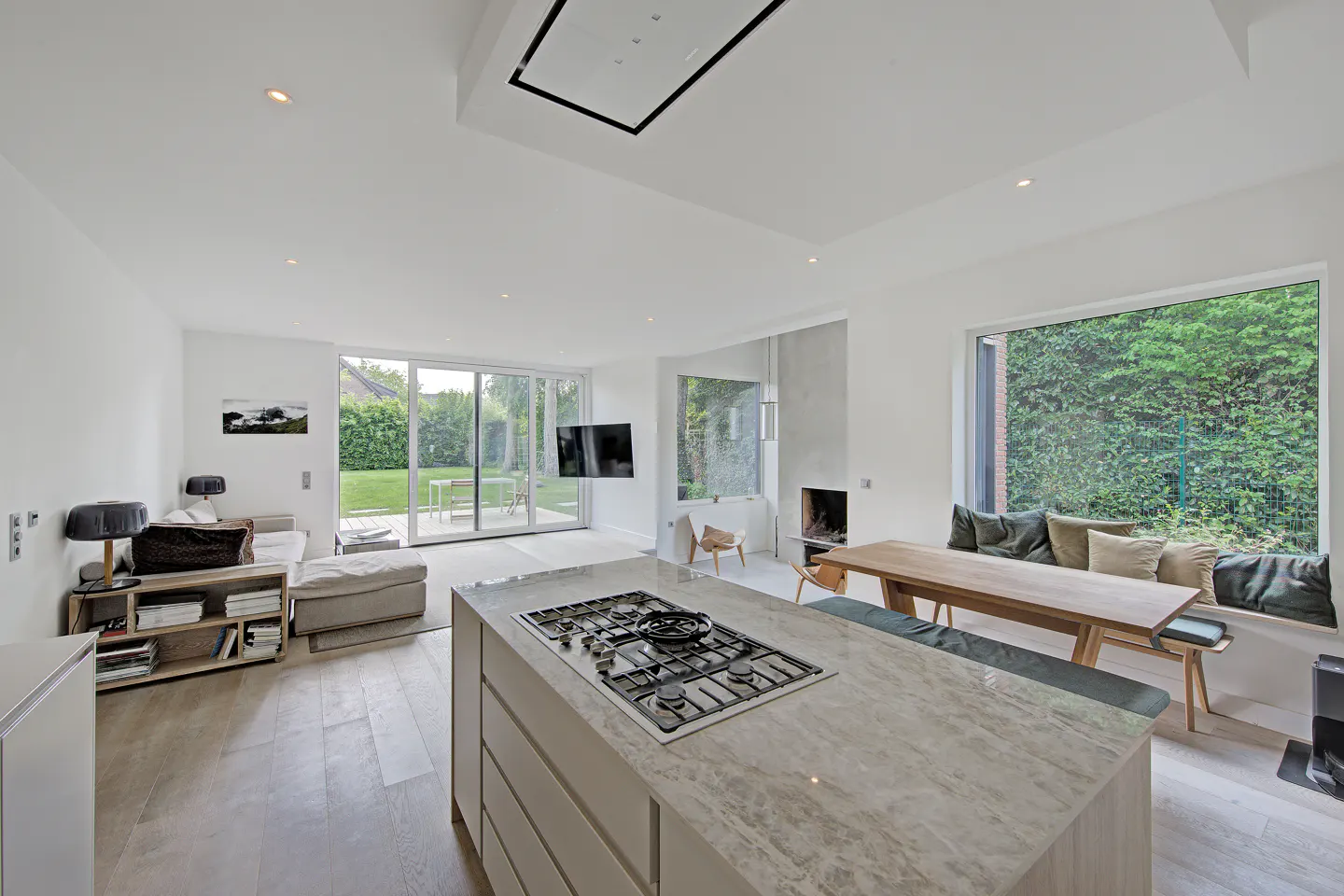 Modern open-plan kitchen and living area with a marble island, wooden dining table, and large windows overlooking a garden.