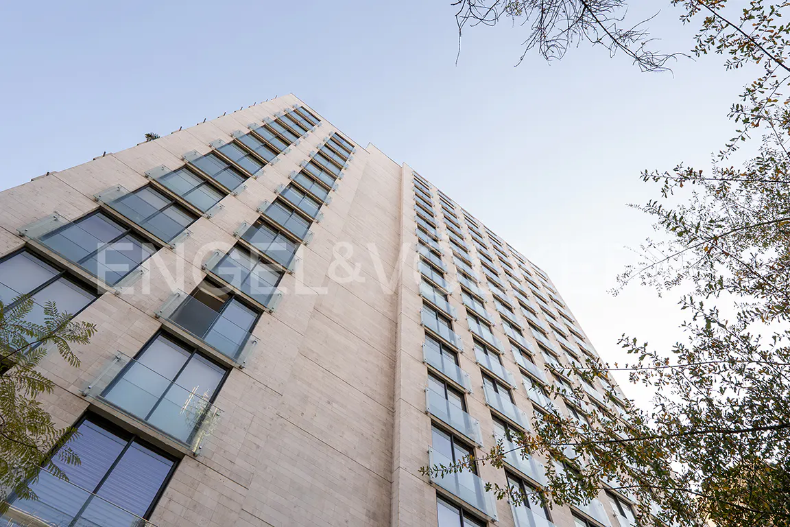 Low-angle shot of a tall, modern apartment building with many windows, against a clear sky and tree branches.