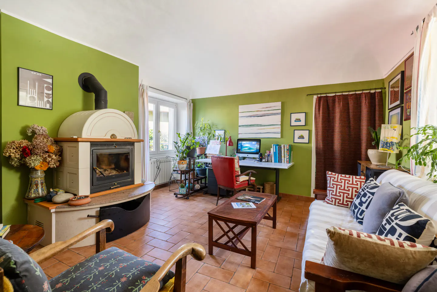 A cozy living room with green walls, terracotta tile floor, a fireplace, desk, sofa, and plants.