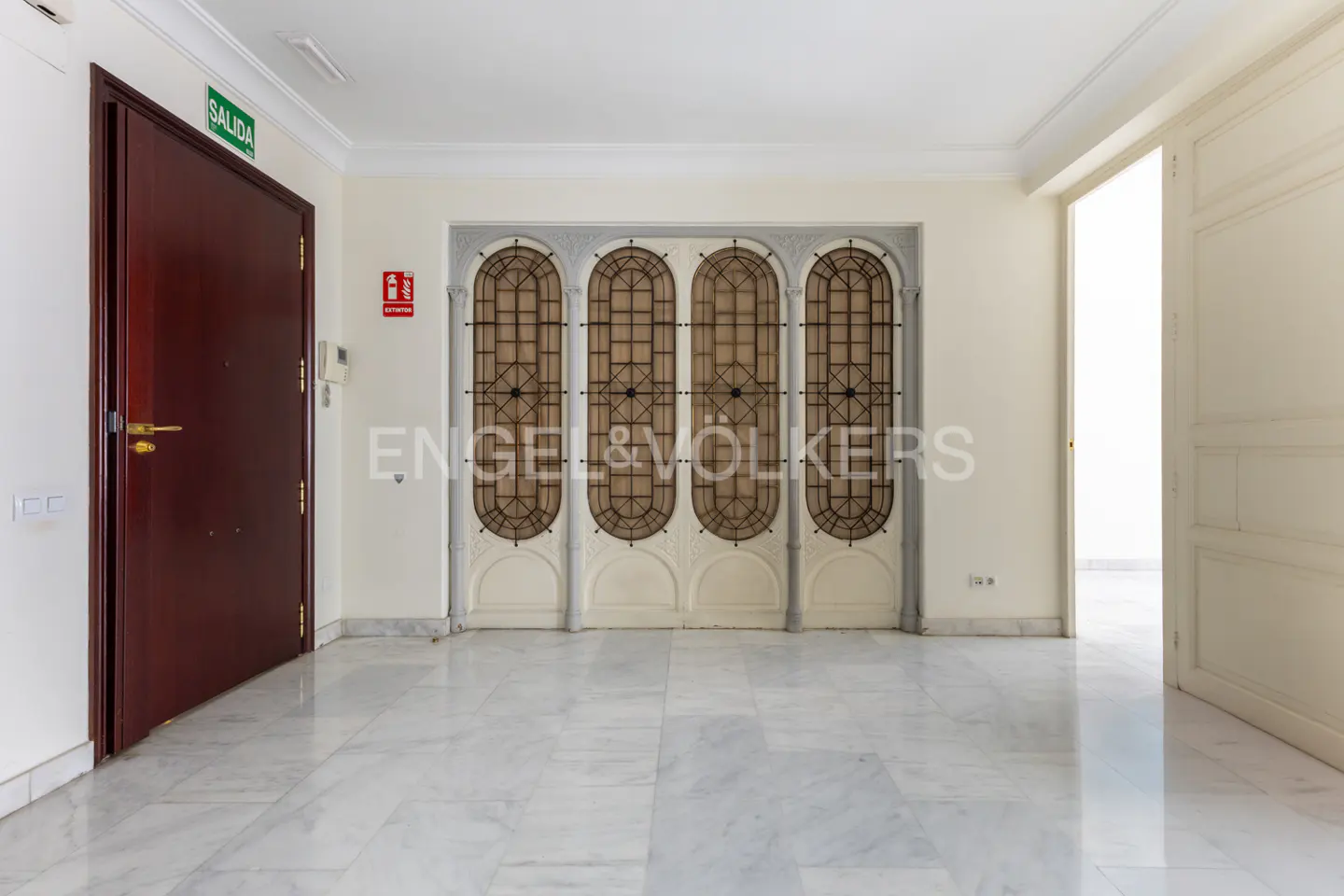 A bright hallway with marble floors, a dark wood door with a green exit sign, and four arched windows.