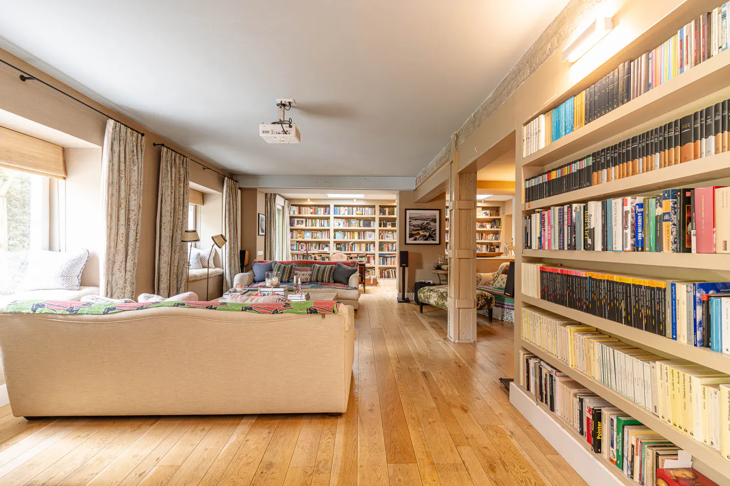 A bright, open living room with hardwood floors and floor-to-ceiling bookshelves filled with books. A beige sofa faces a window seat.