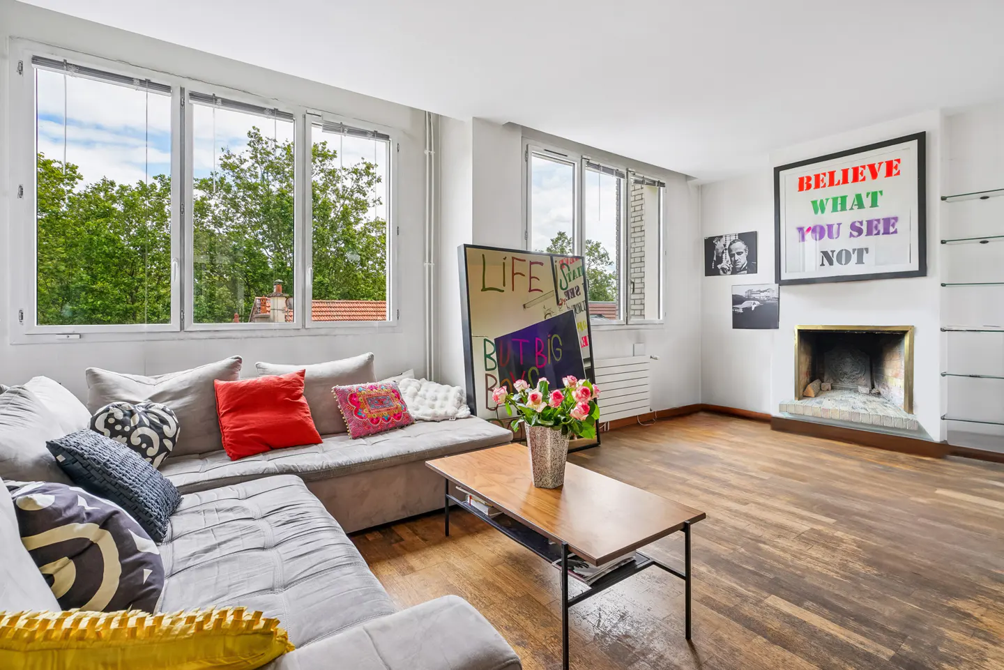 Bright living room with a grey sectional sofa, wooden coffee table, fireplace, and large windows overlooking trees.