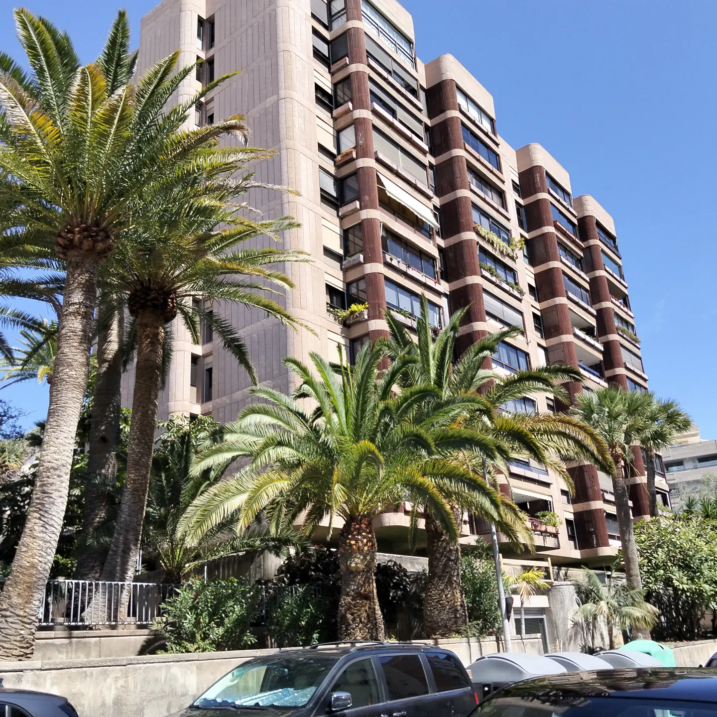 Exterior view of a tall, beige apartment building with balconies, framed by palm trees under a blue sky. Cars are parked in front.