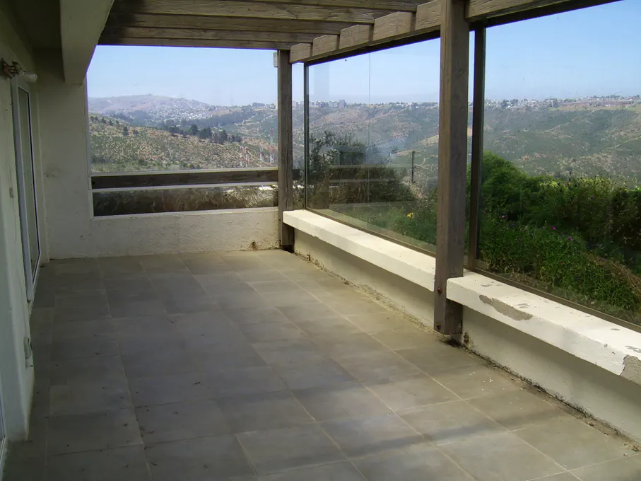 A tiled patio with a wooden pergola overlooks a green hillside and distant city. Glass panels provide a clear view.