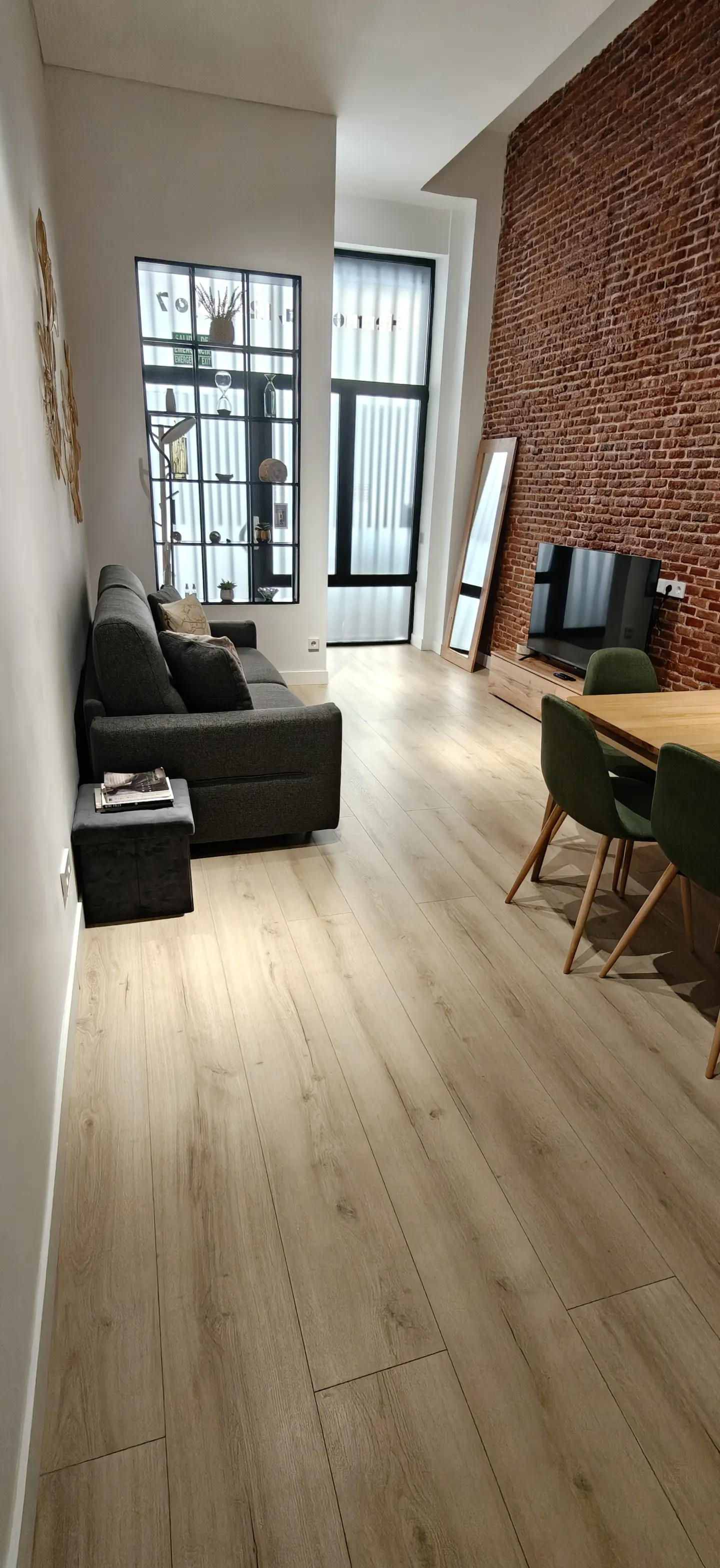 A modern living room with a gray sofa, wooden floors, and a brick wall. A dining table with green chairs is visible.
