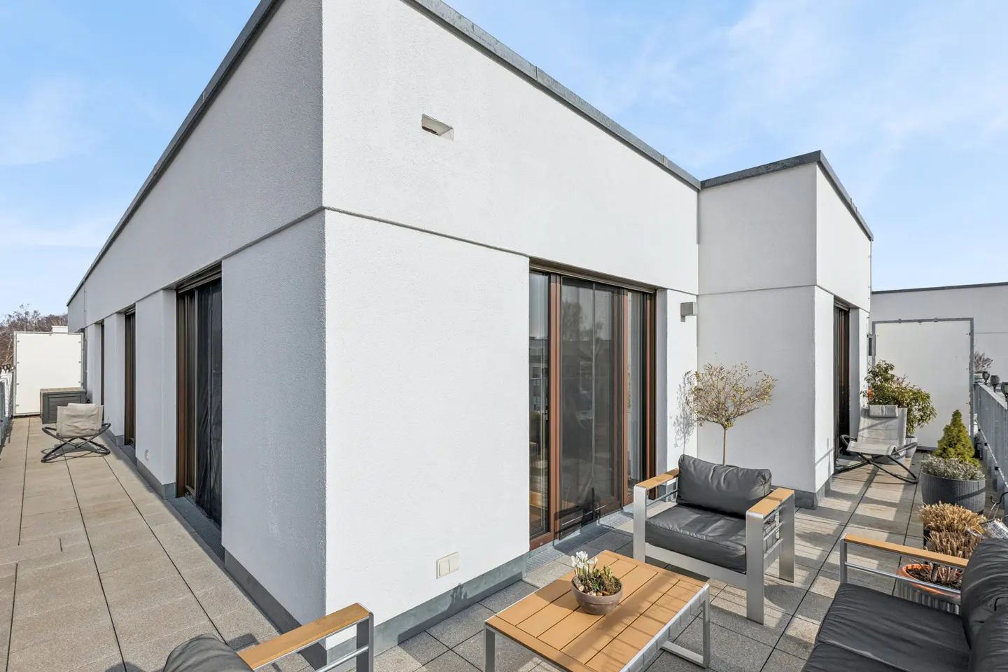 A modern rooftop terrace with gray tile flooring, white walls, and dark-framed sliding glass doors. Outdoor furniture includes chairs, a sofa, and a wooden coffee table.