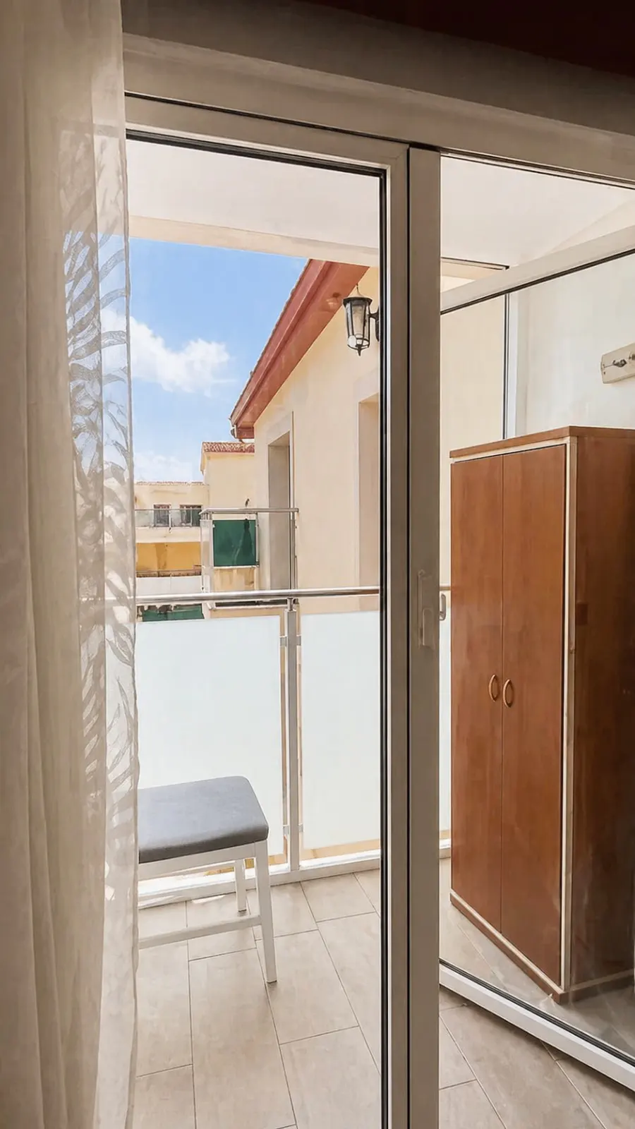 View from a sliding glass door to a balcony with a gray bench and a building in the background. Inside, a brown wardrobe stands.