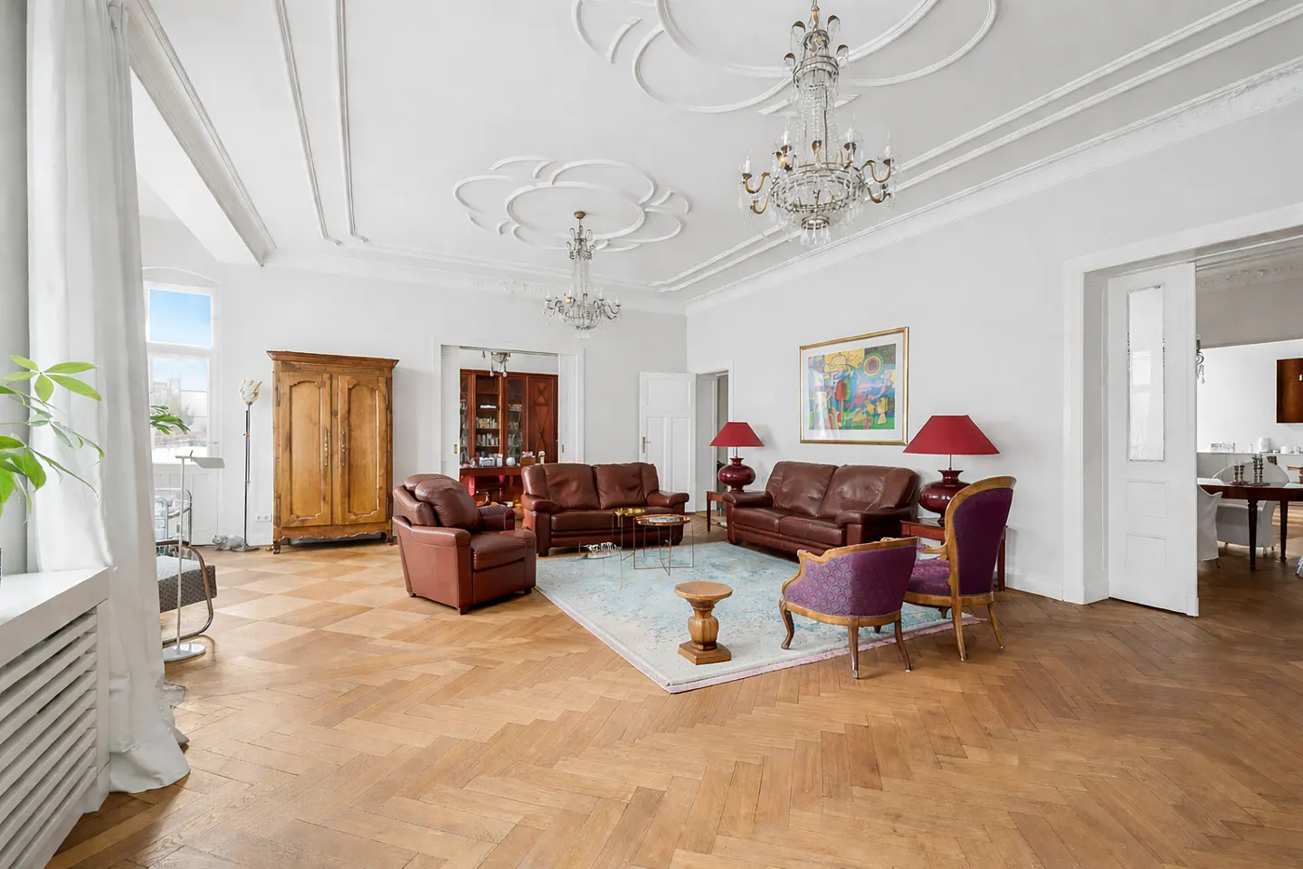 Bright living room with wood floors, white walls, and brown leather sofas. Two chandeliers hang from the ornate ceiling.