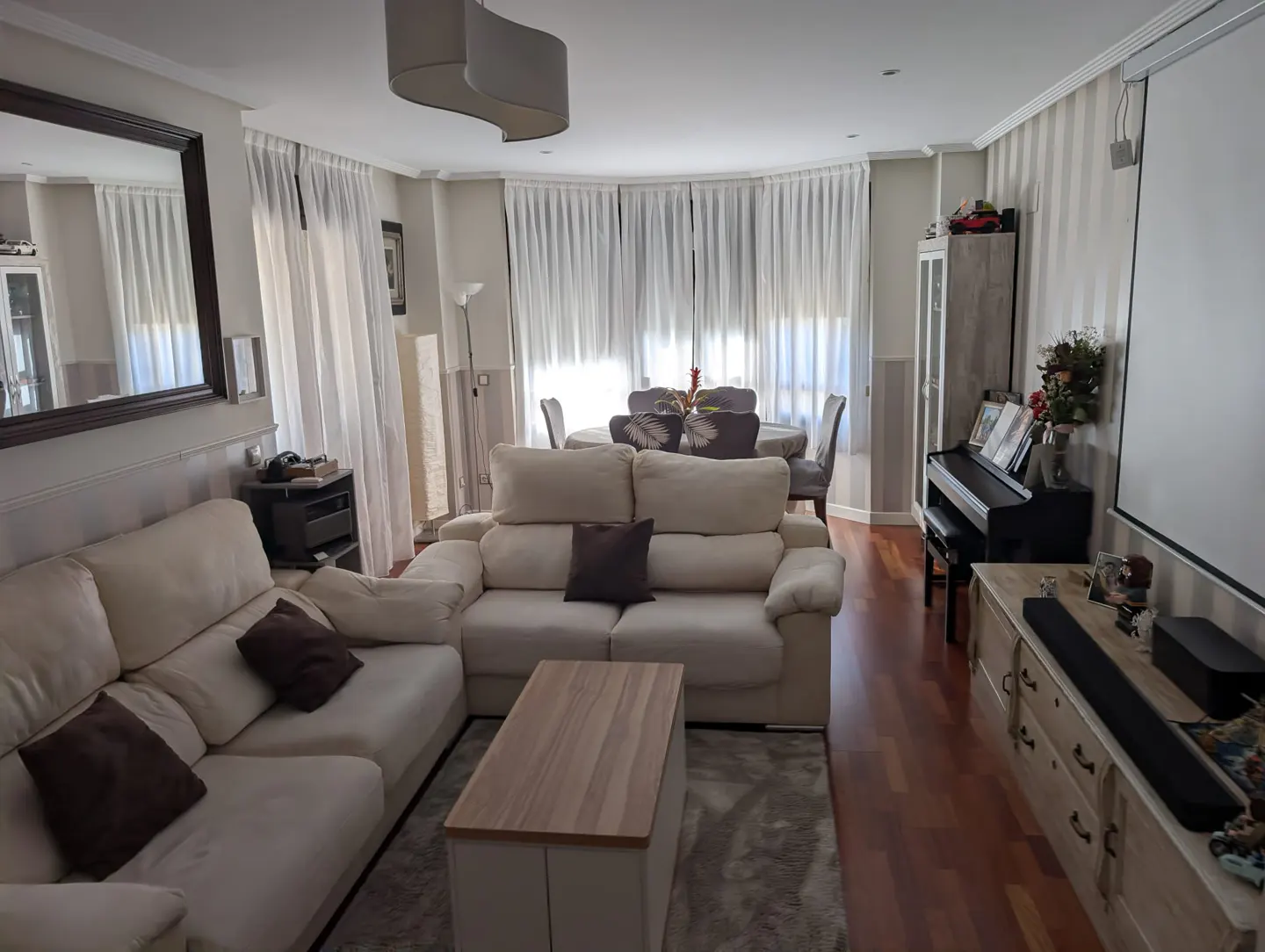 Bright living room with cream sofas, wooden coffee table, piano, and dining area with white curtains and hardwood floors.