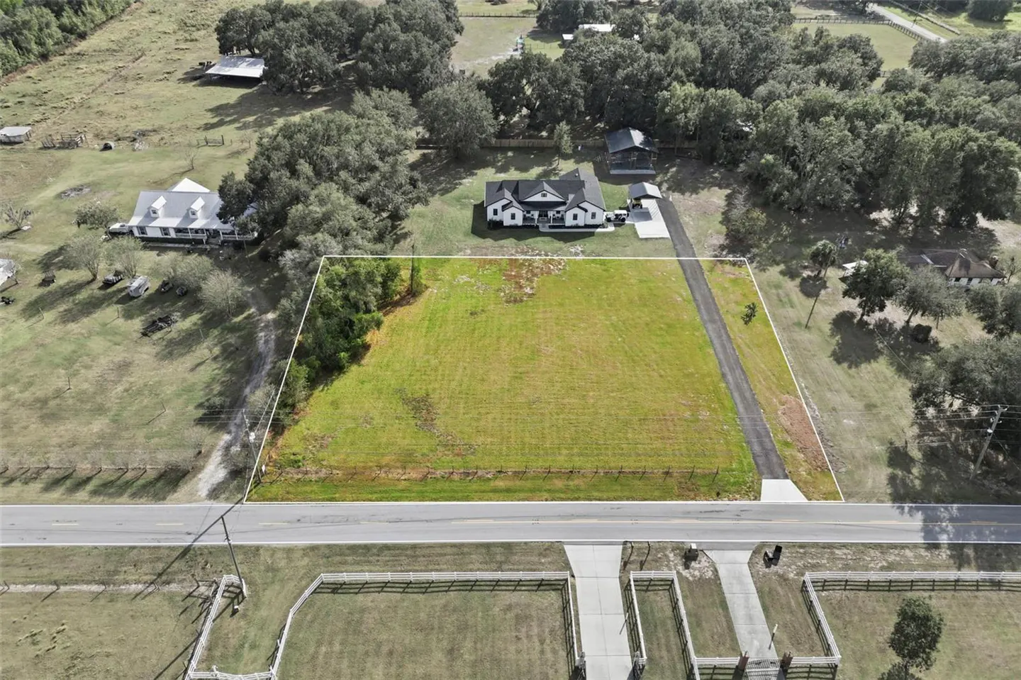 Aerial view of a large, rectangular grassy lot outlined in white, situated next to a paved road and a white farmhouse.