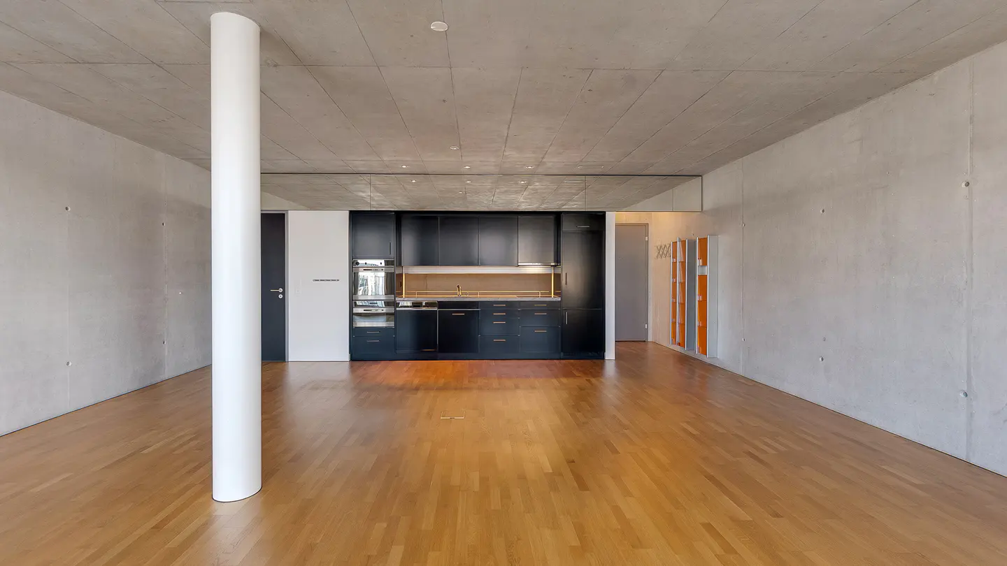 A modern apartment interior with concrete walls, wood floors, and a black kitchen. A white pillar stands in the foreground.