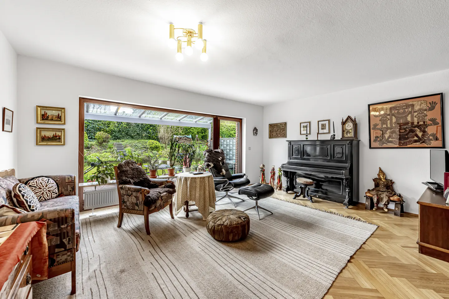 Bright living room with a piano, sofa, chairs, and a large window overlooking a lush green garden.